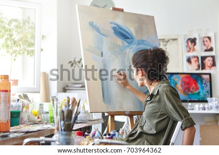 Art, creativity, hobby, job and creative occupation concept. Rear view of busy female artist sitting on chair in front of easel, painting with fingers, using white and blue oil or acrylic paint