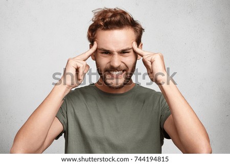 Headshot of young man holds fingers on temples, has bad memory, tries to concentrate and remember details of accident, presses teeth with irritation. Overworked man has headache, feels tired