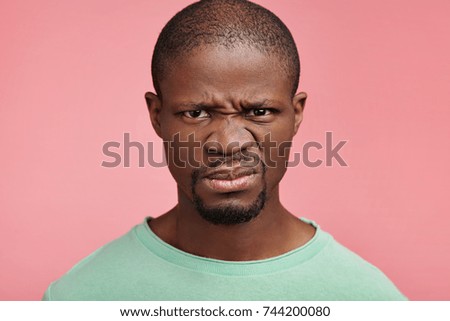 Displeased discontent African American male frowns face, dislikes something, looks with disgusting expression, isolated ovr pink background. Attractive unhappy young man isolated over pink background