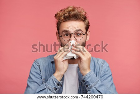 Indoor shot of bearded man has running nose, rubs nose with handkerchief, being ill, caught cold after long walk outdoors on rainy weather. Unhealthy allergic male sneezes isolated over pink wall