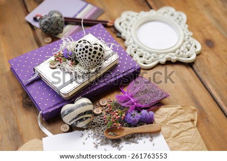 Artistic composition with lavender seeds and flowers next to a purple diary notepad under a white address book and a heart next to a white photo frame, wooden spoon and a sheet of paper on brown wood
