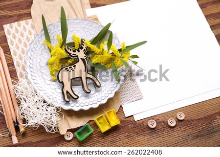 Nature composition with baby deer placed over yellow flowers in rounded white plate over shredded paper, pencils and watercolors with white placeholder paper on the right on top of brown vintage wood