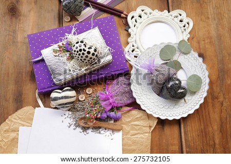 Artistic composition with a purple diary notepad and an organza bag filled with lavender seeds and flowers next to a white rounded platter, notebooks and photo frame over white and brown paper on wood