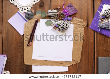 Artistic composition with green leaves, purple organza bag with lavender seeds and flowers next to a purple diary notepad placed next to white photo frame, placeholder sheet of paper over brown wood
