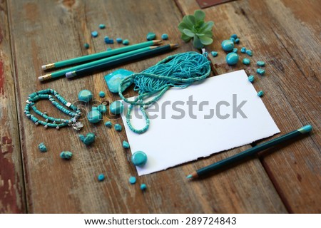Abstract composition from turquoise crystals, stone buddha head and loose jewelry beads next to white placeholder sheet of paper, of succulent green plant flower and pencil over old brown vintage wood