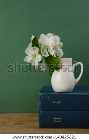 A stack of books and bird cherry branch in a jug on the wooden table