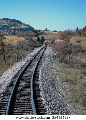 A narrow-gauge railroad track heads toward the horizon through pasture land and past a mountain covered with aspen trees in bloom