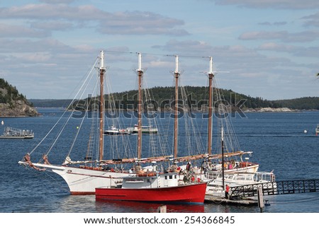 A large white sailboat, or schooner, and red fishing boat are anchored in the ocean of Bar Harbor, Maine with islands in the distance.