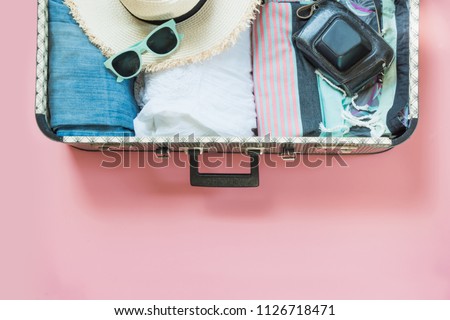 Open suitcase with female clothes for trip on pastel pink background. Top view with copy space. Summer concept travel.