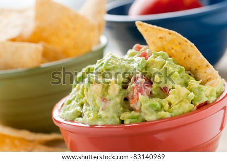 Guacamole and Chips - A studio shot of homemade guacamole in a red bowl., tortilla chips in a green bowl and a tomato in a blue bowl.