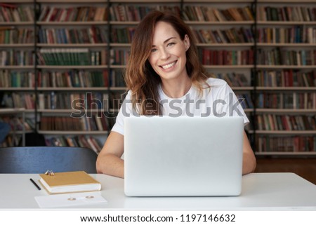 Woman entrepreneur working on laptop at co-working office or library, looks to camera, smiling, bookshelves. Knowledge and self-development.