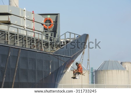 Port of Amsterdam, Noord-Holland/Netherlands - August 26-08-2015 - Sailor is painting the ships name Wilson Vivero on the bow. The sailor is sitting on a small wooden bench. factory at the background.