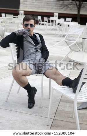 cheeky  young man in fashionable glasses wearing grey shorts with hairy legs sitting on chair at street cafe