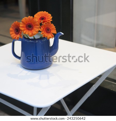 orange gerbera flower with water drops in blue tea-pot on the white table as a decoration of the street cafe