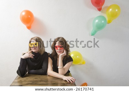two  woman wearing funny glasses sitting at the table. Wall decorated balloons.