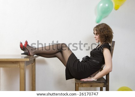 lonely girl in black dress sitting on chair in empty room putting her legs down table. Woman wearing red shoes. After holiday.