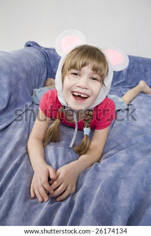 Little happy giggle girl wearing cap with mouse`s ears lying on sofa. On her seven birthday.