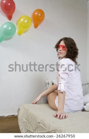 young curl strange woman wearing funny glasses sitting on the bed at wall. Wall decorated balloons.