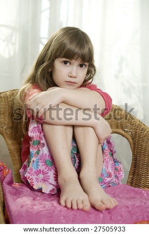 Little cute dreaming girl sitting on chair at window hugging her knees