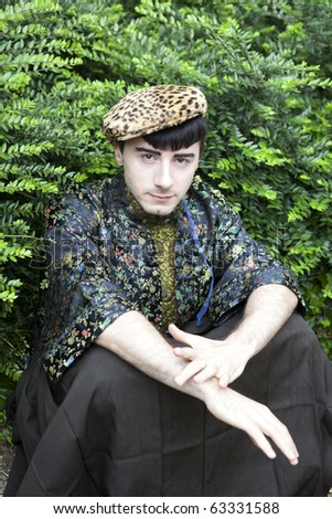serious man in leopard cap wearing kimono sitting opposite green bush looking in camera