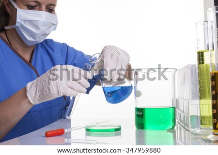 lab technician in scrubs and mask and gloves holding a test tube and beaker shot front on at a desk on a white background landscape