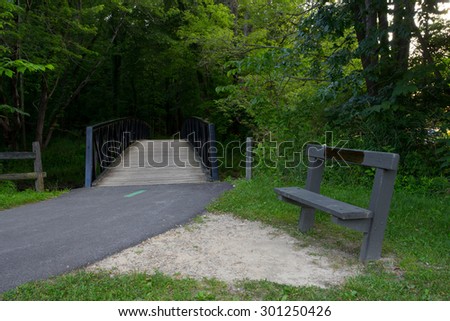 Park bench near bridge. Outdoor background shot showing a bench near an outdoor park trail and walking bridge. Bench is on right of trail.