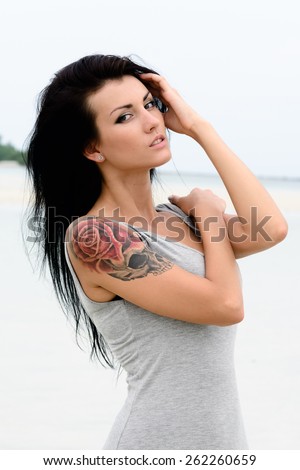 Cute young sexy girl in grey dress posing on the beach, blue sea background, brutal tattoo