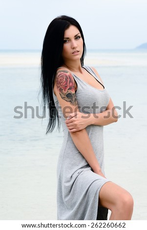 Cute young sexy girl in grey dress posing on the beach, blue sea background, brutal tattoo