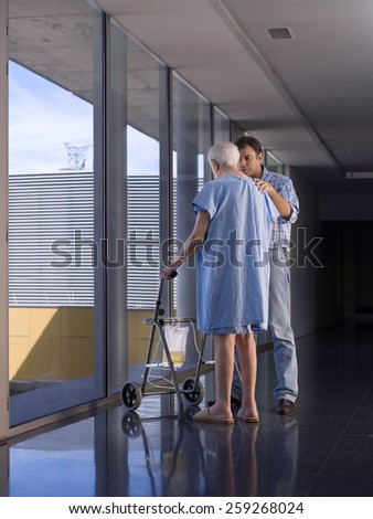 Elderly person looking out a window of a hospital, with the help of a walker and with his son