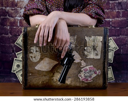 Suitcase full of money, and above it, the hands of a woman holding a gun