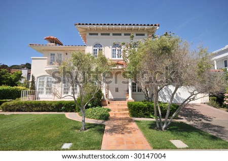 House with trees on each side of the walkway to the front double doors. Blue sky. Two levels with grass yard, trees and shrubs.