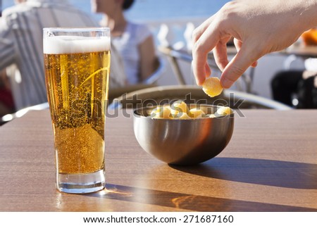 A hand picking snacks from a bowl and a beer in a table of a terrace near the sea in a sunny summer day.
