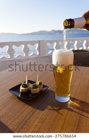 Pouring cold beer from the bottle in a glass and a plate of green peppers in the table of a terrace next to the sea in Spain.