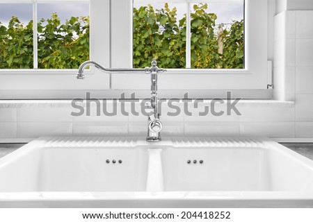 Retro water faucet on new empty white kitchen sink with view on vineyard through the window in background.