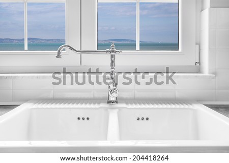 Retro water faucet on new empty white kitchen sink with view on sea through the window in background.