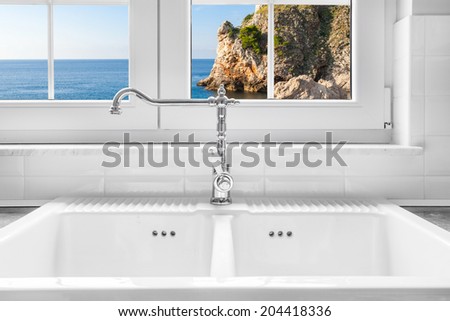Retro water faucet on new empty white kitchen sink with view on sea rock through the window in background.