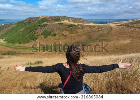 Young woman with arms wide open enjoying the view on hills in Holyrood Park in Edinburgh, Scotland. Park\'s highest point Arthur\'s seat is popular destination for hiking and enjoying nature.