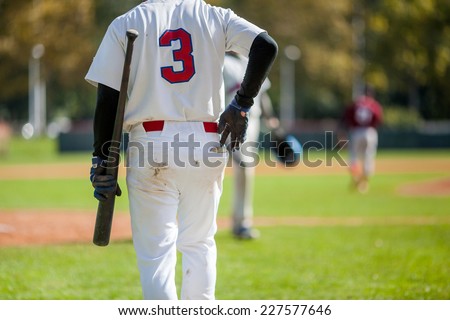 Closeup from the back of baseball player in white jersey  holding bat with the left hand and reaching for ball from back pocket with the right hand.