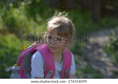 The girl came home from school,sitting with a backpack in the sun blonde, beautiful with pink big backpack behind the green background, outdoors