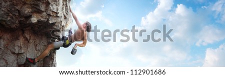 Young man climbing vertical wall with belay with blue cloudy sky on the background
