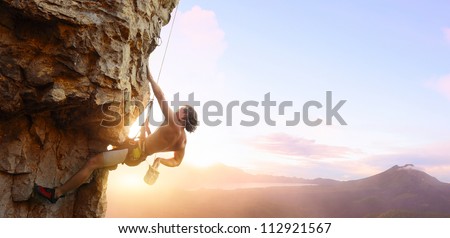 Young Man Climbing Vertical Wall With Belay With Sunrise Valley On The Background
