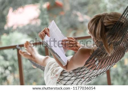 Woman in summer dress lies in the hammock in a garden and reads the book