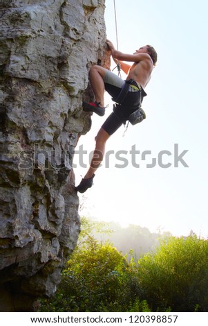 Young man climbing vertical natural rocky wall with clear sky on the background