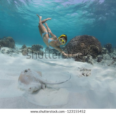Underwater shoot of a woman swimming over sandy sea bottom with some corals and watching spotted ray