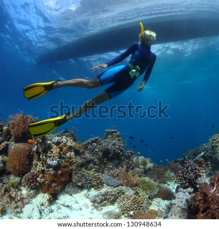 Underwater Shoot Of A Young Lady Snorkeling And Diving On A Breath Hold Over Vivid Coral Reef Of Tropical Island With Boat Silhouette On Surface