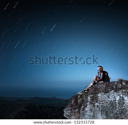 Tourist with backpack relaxing on top of a mountain at bright night with stars in a sky