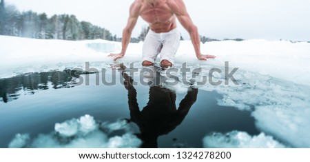 Young man with lean muscular body going to swim in the cold winter water with ice floating on the surface and forest on the background. Tilt shift effect applied, focus on the floating ice pieces