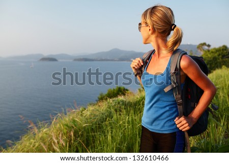 Young lady with backpack standing on a lush tropical meadow and looking to horizon