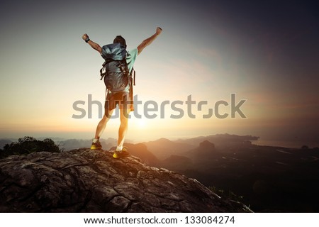 Hiker with backpack standing on top of a mountain with raised hands and enjoying sunrise