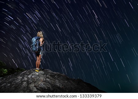 Hiker with backpack standing on top of a mountain with star trails on the background (real stars)